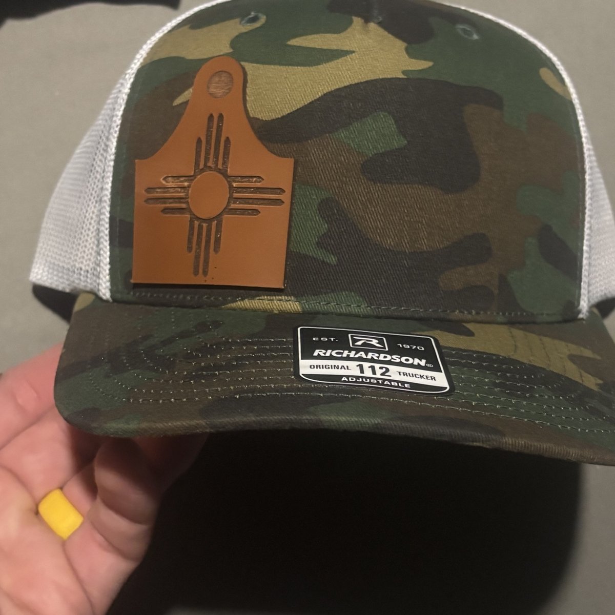 Camo Richardson 112 hat with a brown leather ear-tag patch with a Zia symbol, by KB Custom Creations