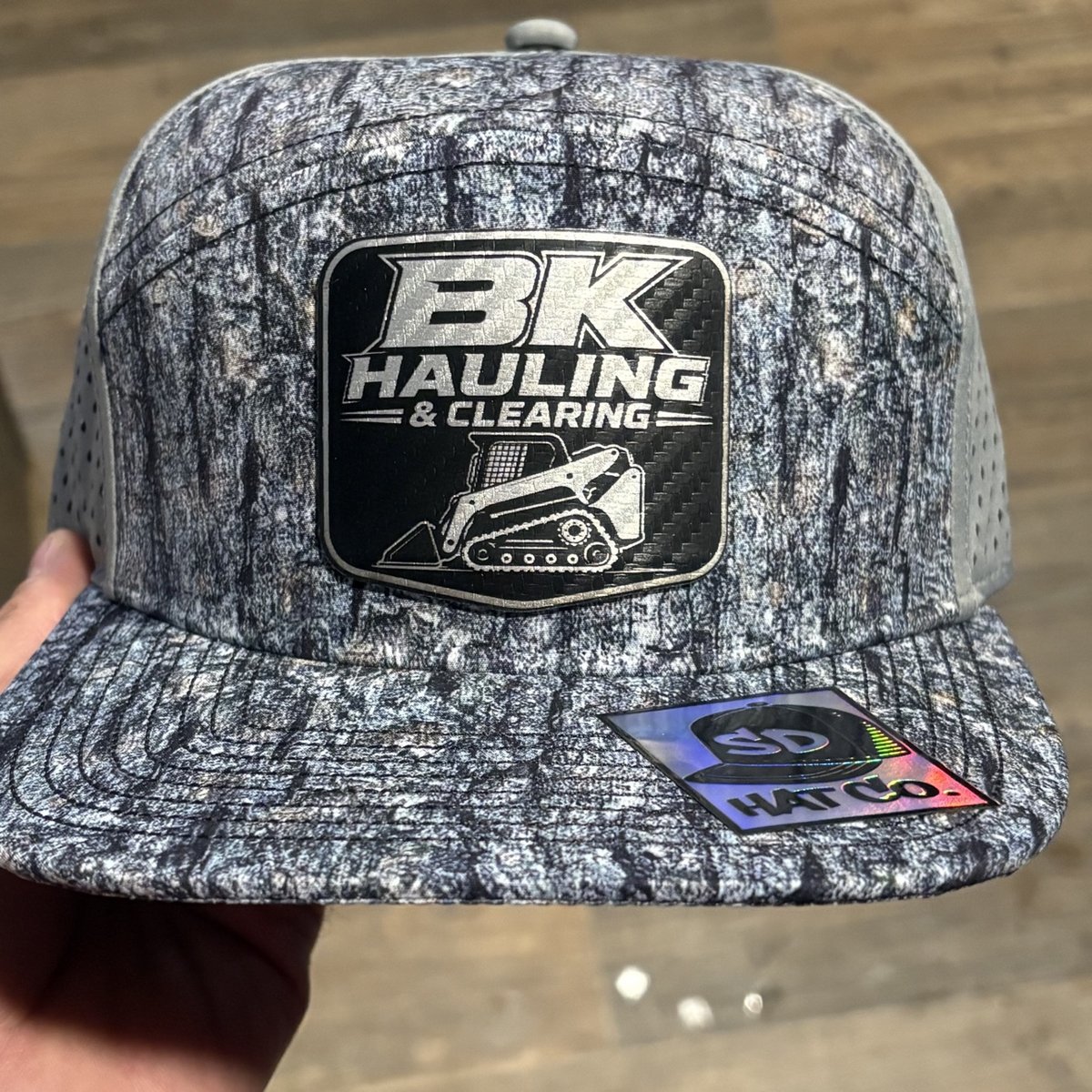 Bark-camo hat with a black BK Hauling and Clearing skid-steer patch, by KB Custom Creations