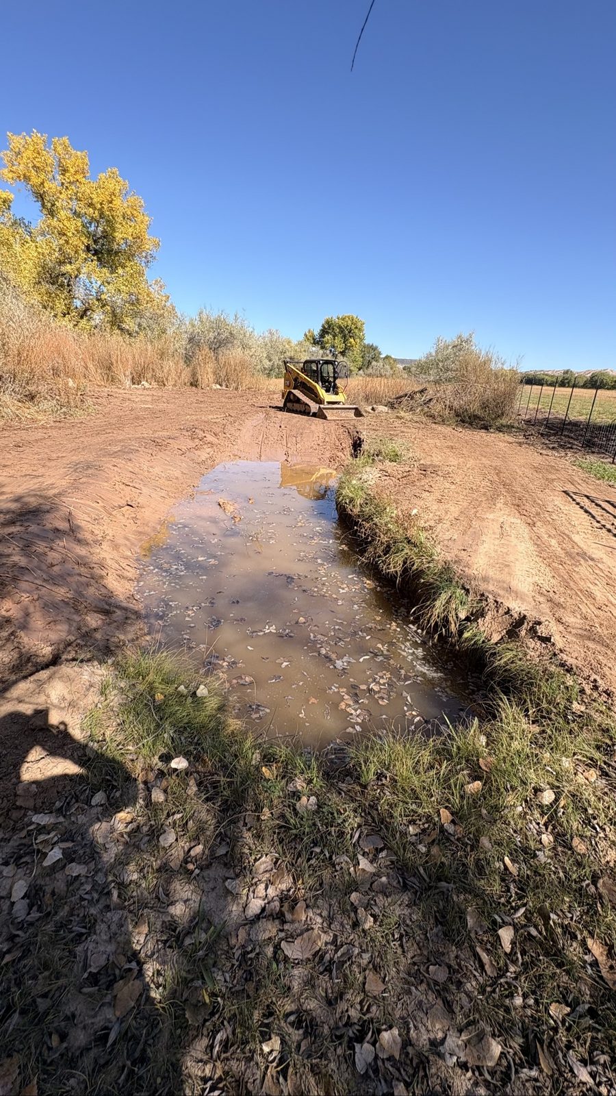 Wide straight-on view of the finished drainage ditch after BK Hauling & Clearing grading — both banks cut smooth, CAT skid steer at the far end