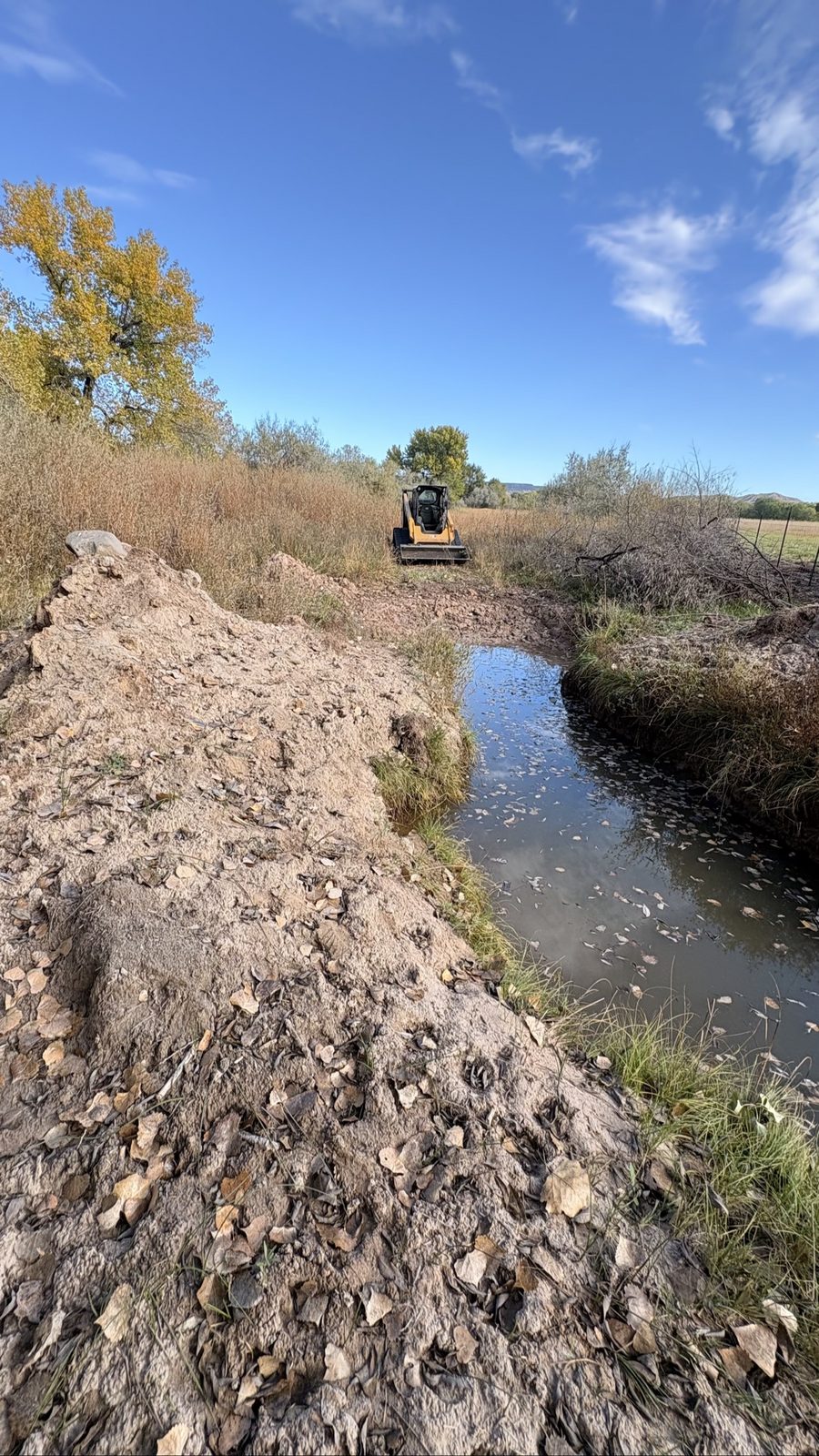 Low view along the rough, ungraded bank of the drainage ditch before BK Hauling & Clearing work, CAT skid steer waiting at the far end