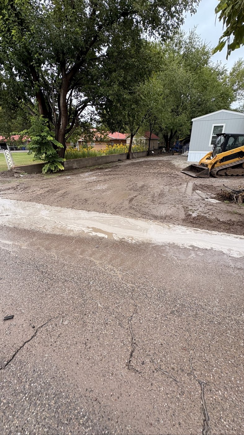Rainy-day driveway job in progress — mud and water sheeting across the lot toward the street, CAT skid steer on site